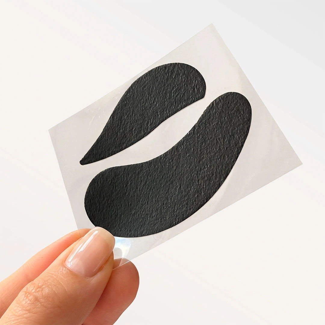 A hand holding a clear backing sheet with a pair of black bio gel upper and lower eye pads.